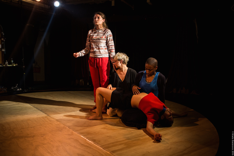 Dancers in a tablau in the center of the platform. A dancer in red pants stands. While another drapes over another dancer's lap.